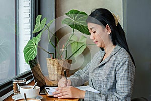 Asian business woman trading portfolio sitting at the table by the window In hand holding pen and checking document lying on table