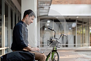 Asian businessman with bicycle using laptop and sitting outside the office building. man commuting on bike go to work