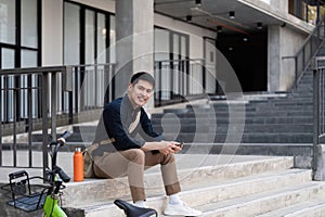 Asian businessman with bicycle using smartphone and sitting outside the office building. man commuting on bike go to