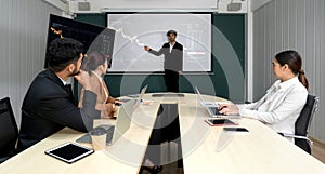 Asian businessman in black suit pointing at multicolor chart on presentation monitor. Business executives team meeting at night in