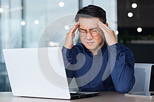 Asian businessman boss sick has severe headache, man in glasses sitting at desk using laptop at work inside office