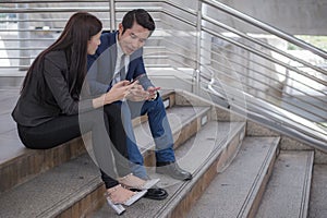 Asian businessman and businesswoman sit on the staircase and using telephone to surf internet for shopping online.