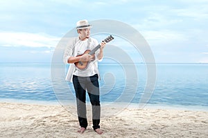 Asian businessman in hat and white clothes playing guitar on the beach