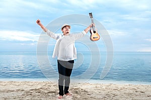 Asian businessman in hat and white clothes raised both hands while holding guitar on the beach