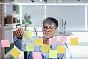 Asian businessman looking and writing on sticky note for brainstorming ideas on clear whtieboard at modern office. analyze and