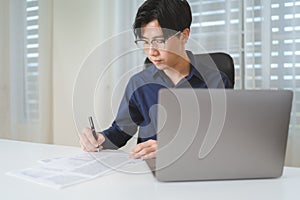 Asian businessman, male businessperson writing on paperwork with computer laptop on desk at office. Professional entrepreneur