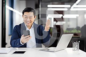 Asian businessman in office suit smiling and waving hello during a video call on smartphone