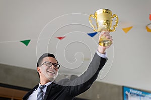 Asian businessman raising up hand and holding a golden trophy cup to cheerful and celebrated his successful in mission.