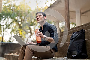 Asian businessman with reusable eco friendly ecological cup using laptop and sitting outside the office building. Eco