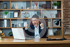 Asian businessman shouts with joy and holds hands up, celebrates victory, man works at desk in office, looks at laptop screen