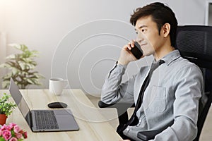 Asian businessman sitting at desk discussing business deal on mobile phone There are laptops and coffee mugs on the wooden table.