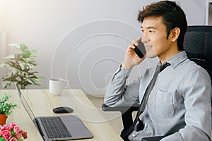 Asian businessman sitting at desk discussing business deal on mobile phone There are laptops and coffee mugs on the wooden table.