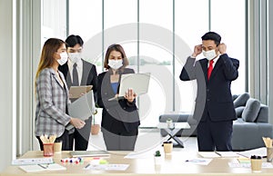 Asian businessman in suit waring a mask, while walking into a group of colleagues. The team is preparing for the meeting. Concept
