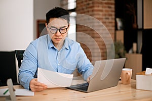 Asian businessman working with documents and laptop at home office, checking financial papers, sitting at workplace