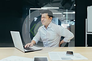 Asian businessman working in office, having severe back pain, overtired worker working with laptop photo