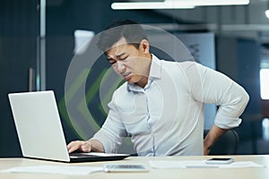 Asian businessman working in office, having severe back pain, overtired worker working with laptop photo