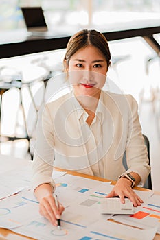 Asian businesswoman analyzing a report pointing to a graph with a pen, laptop, and calculator. In the online marketing concept of
