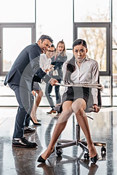 Asian businesswoman bound with rope on chair and multicultural business team pulling her