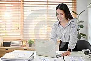 Asian businesswoman leaning on table, using her laptop, working on her business assignment