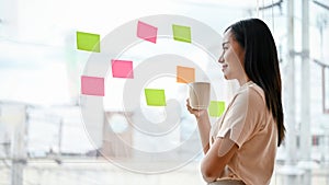 An Asian businesswoman sipping coffee and looking at the sticky notes on a glass wall