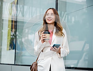 Asian businesswoman with smartphone and cup coffee standing against street