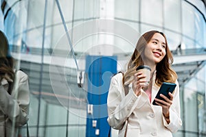 Asian businesswoman with smartphone and cup coffee standing against street building near office