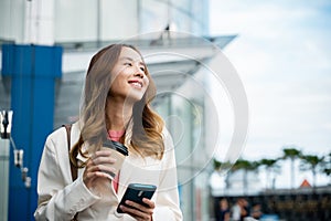 Asian businesswoman with smartphone and cup coffee standing against street building near office