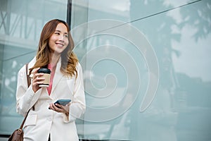 Asian businesswoman with smartphone and cup coffee standing against street building near office