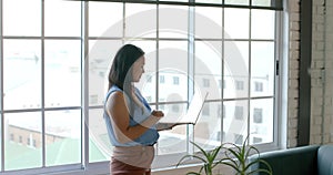 Asian businesswoman stands by a window in an office, holding a laptop