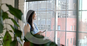 Asian businesswoman stands by a window in an office setting, holding a tablet, with copy space