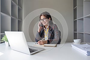 Asian businesswoman talking on mobile phone working on laptop in modern office. Happy business woman talking on mobile