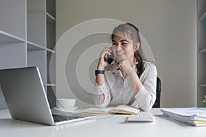 Asian businesswoman talking on mobile phone working on laptop in modern office. Happy business woman talking on mobile