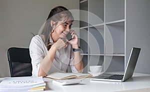 Asian businesswoman talking on mobile phone working on laptop in modern office. Happy business woman talking on mobile