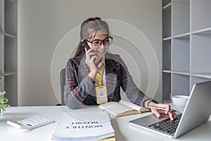 Asian businesswoman talking on mobile phone working on laptop in modern office. Happy business woman talking on mobile