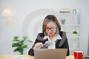 Asian businesswoman wearing suit and glasses holding magnifying glass and working laptop. Seaching browsing. Entrepreneur woman or