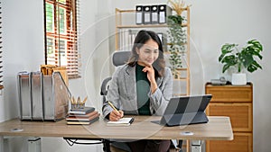 Asian businesswoman working at her desk, hand on chin, lookin at tablet screen