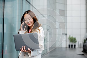 Asian businesswoman working on laptop and talking on cell phone at front building near office