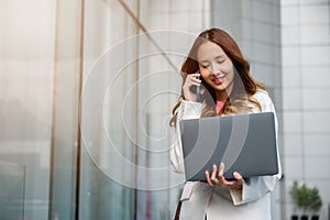 Asian businesswoman working on laptop and talking on cell phone at front building near office