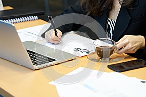 Asian businesswoman writing sales report with black pen On the desk with a notebook computer. And sales report paper. using as bac
