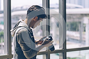 Asian Camera man holding camera sitting near big windows glass of building and looking Play back Pictures from camera