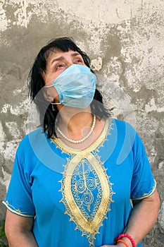 Asian or caucasian ethnicity mature woman in blue dress with medical mask on his face to protect COVID-19, looking up against