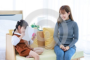 Asian cheerful happy young pretty female mother nanny babysitter in casual outfit sitting on sofa smiling hugging little cute daug