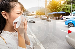 Asian child girl blowing nose in paper handkerchief,female person sneeze in a tissue in city street,toxic fumes from car,bad smell