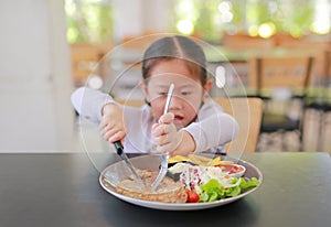 Asian child girl eating Pork steak and vegetable salad on the table with holding knife and fork. Children having breakfast