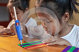 Asian child girl glueing colored ice cream sticks by hot melt electrical glue gun.