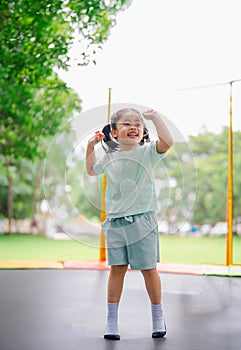 Asian child girl is jumping on trampoline on playground background. Happy laughing kid outdoors in the yard on summer vacation.
