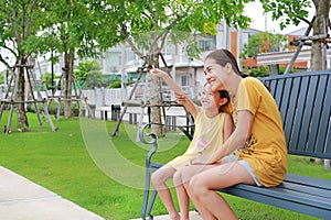 Asian child girl point to looking something with mother in summer park. Mum and daughter relaxing sitting on bench in garden