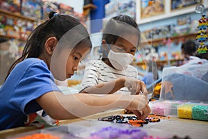 Asian child girl and sister wearing protection mask making fusible beads thermo mosaic in pegboard that is a toy to develops the