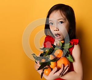 Asian child in red blouse. Looking wondered, holding an armful of tangerines and oranges, posing on orange background. Close up