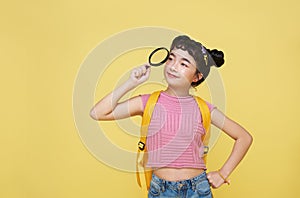 Asian child schoolgirl stood in casual wear looking at camera through magnifier while standing bright yellow background